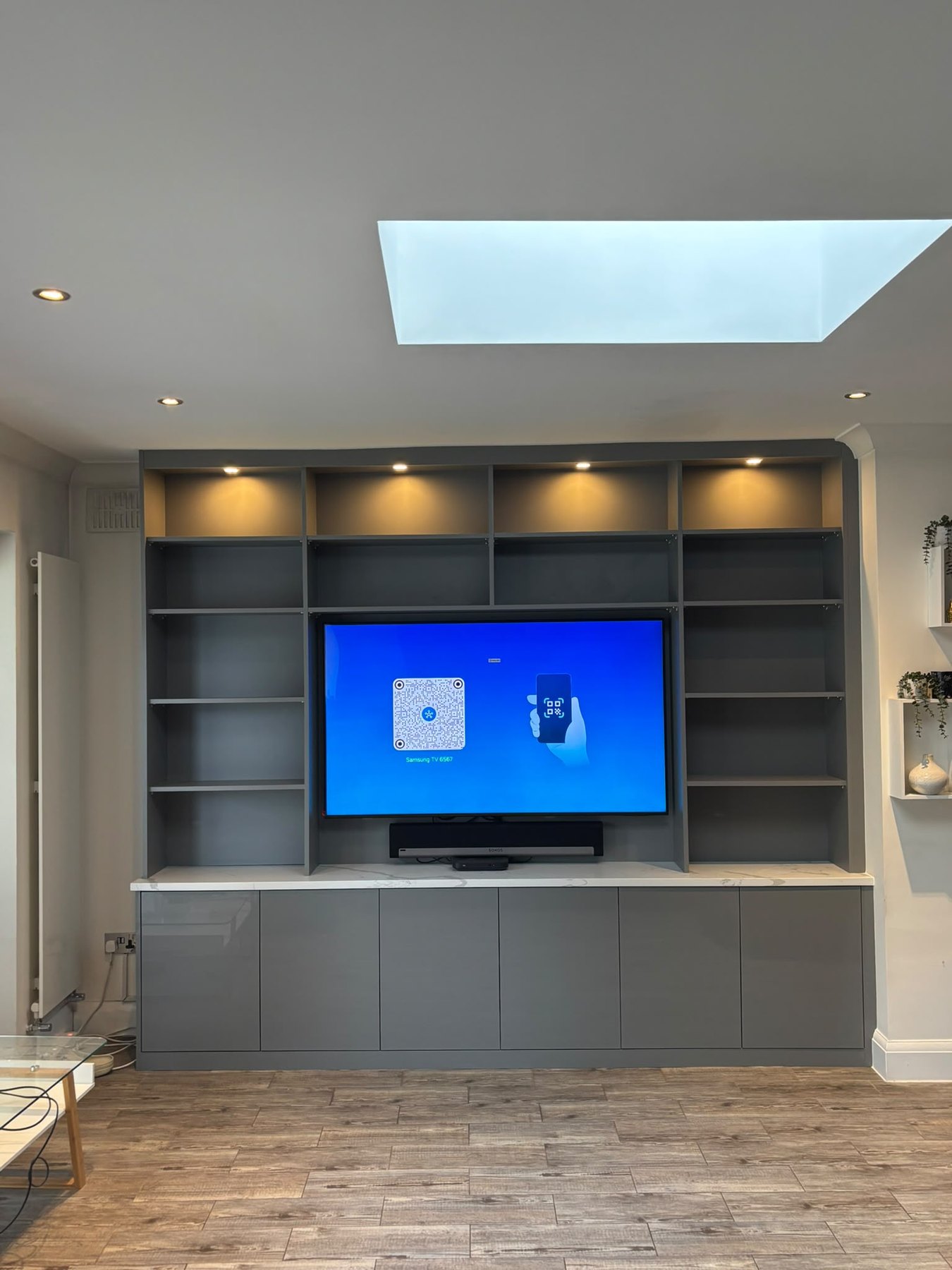 Grey fitted media wall with a recessed television, open shelving lit by integrated LED downlights, a stone worktop and gloss cupboards below