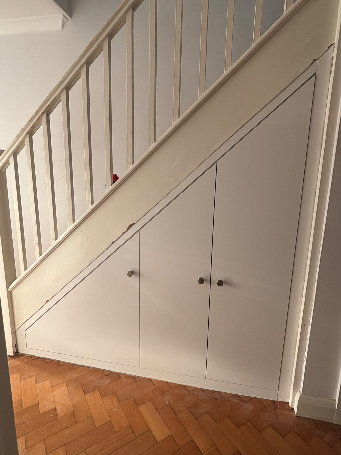 Painted understairs storage with a run of cupboard doors following the line of the staircase, over a parquet floor