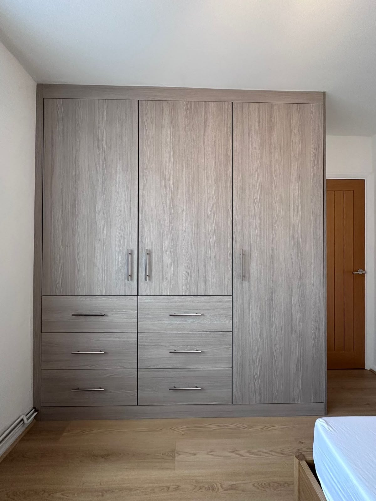 Full-height fitted wardrobe in grey oak with three doors and six drawers, built wall to wall in a bedroom, Feltham